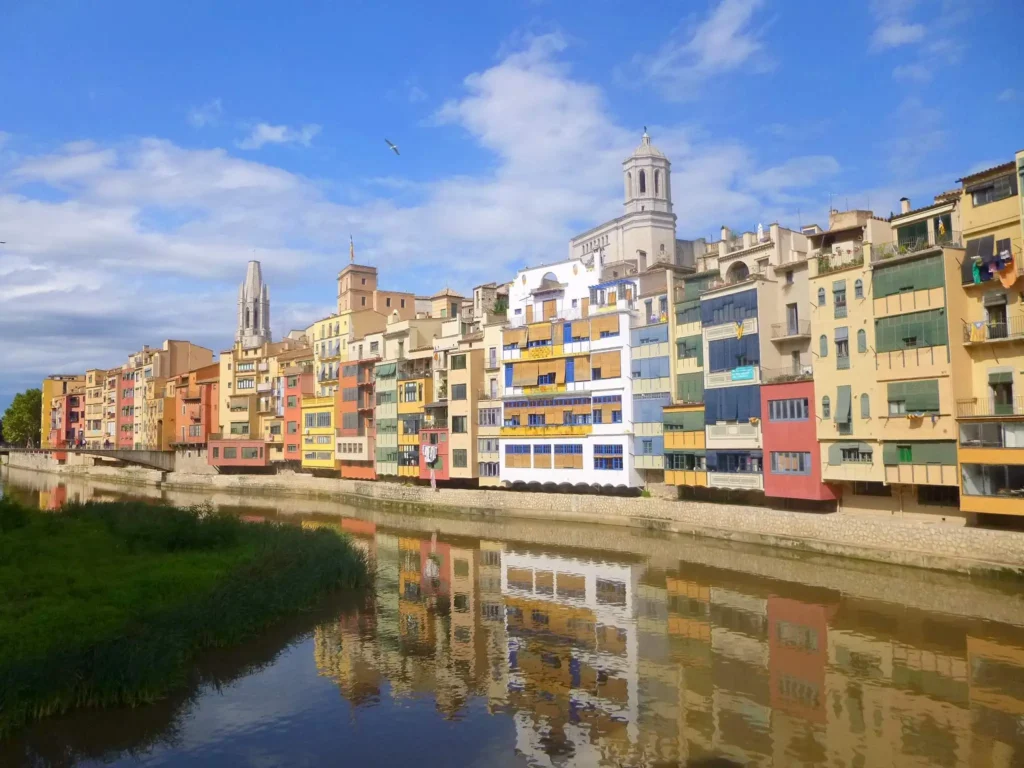 Fachadas de colores del casco antiguo de Girona reflejadas en el río Onyar, con el cielo azul y torres históricas al fondo.