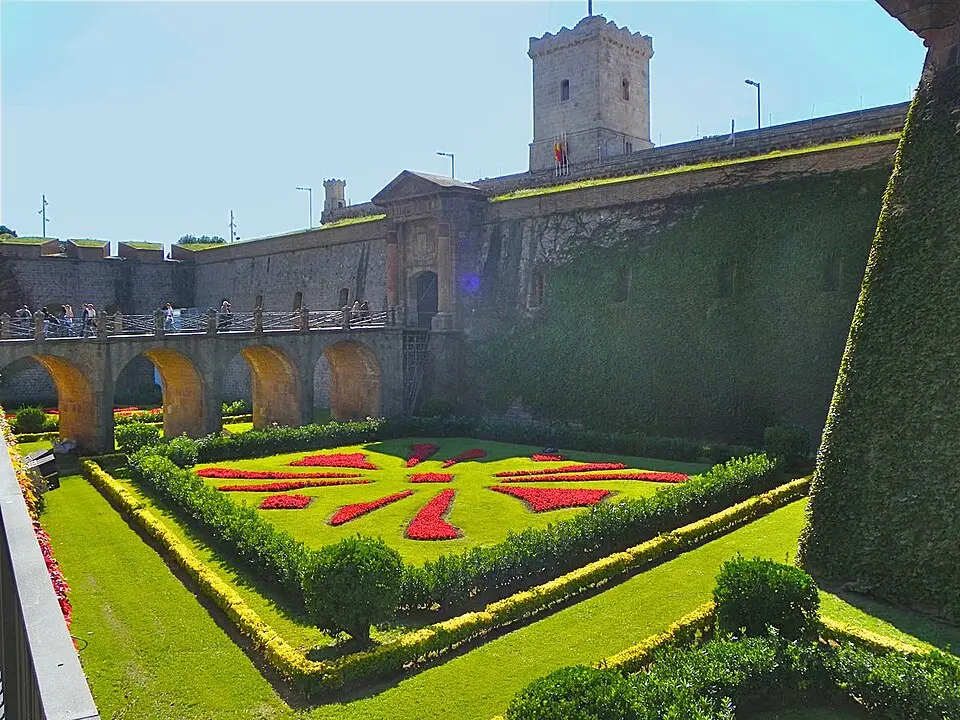 Vue du Castell de Montjuïc à Barcelone avec le pont d’accès et des jardins géométriques ornés de fleurs rouges, entourés de remparts historiques.