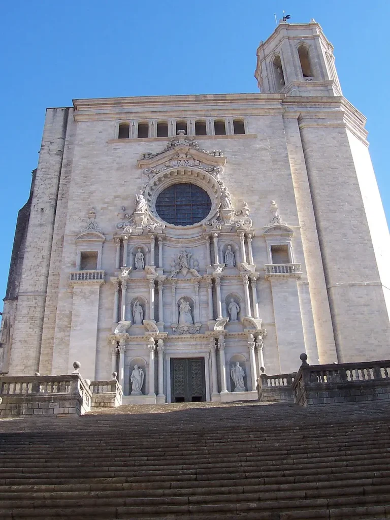 Fachada barroca de la Catedral de Girona vista desde la escalinata, con esculturas y gran rosetón bajo un cielo azul despejado.