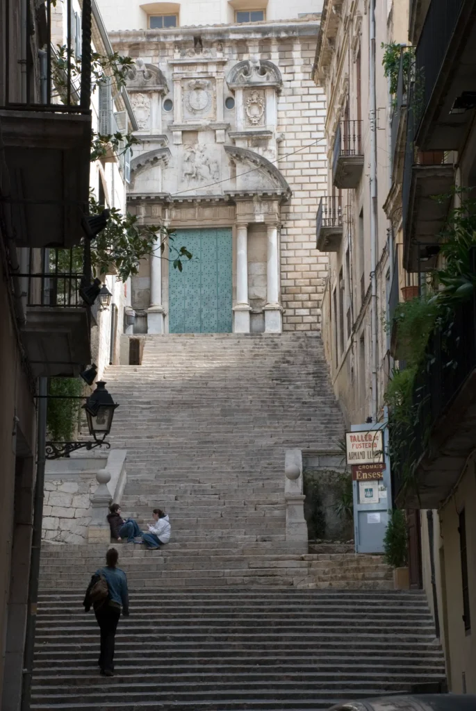 Vista de las escaleras de Sant Martí Sacosta en el casco antiguo de Girona, con una fachada histórica y una gran puerta verde al fondo.