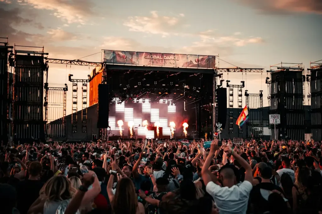 Imagen de un festival de música al aire libre con un gran escenario iluminado, llamas en el escenario y público multitudinario disfrutando del concierto al atardecer