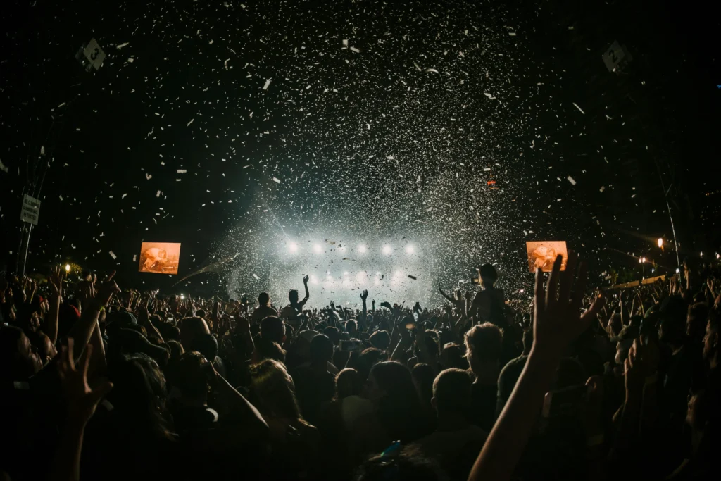 Imagen de un gran concierto nocturno con multitud de personas levantando las manos frente a un escenario iluminado, confeti en el aire y ambiente festivo de un festival de música