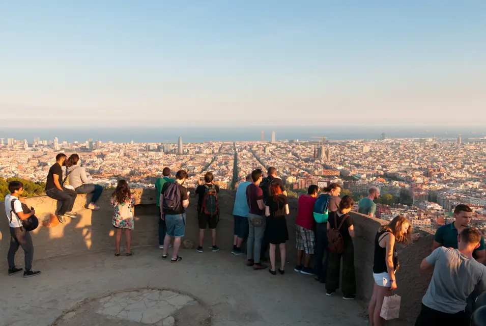 Personas disfrutando de las vistas panorámicas de Barcelona desde los Búnkers del Carmel al atardecer, con la Sagrada Familia y el mar al fondo.