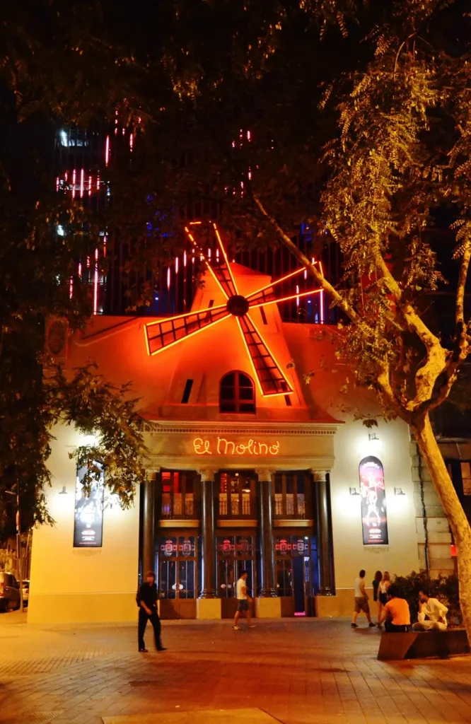 Fachada de El Molino en Barcelona iluminada con luces rojas por la noche, con su icónico molino y personas paseando por la avenida Paral·lel