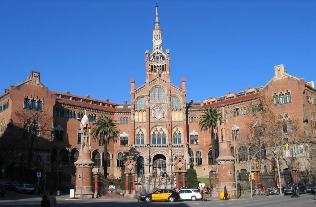 Fachada principal del Hospital de Sant Pau en Barcelona, un emblemático edificio modernista con torre central, detalles arquitectónicos y palmeras en la entrada.