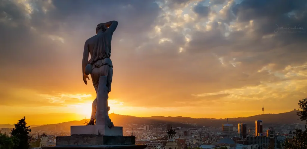 Estatua en Montjuïc con vistas panorámicas de Barcelona al atardecer, con el cielo iluminado en tonos cálidos y la ciudad al fondo