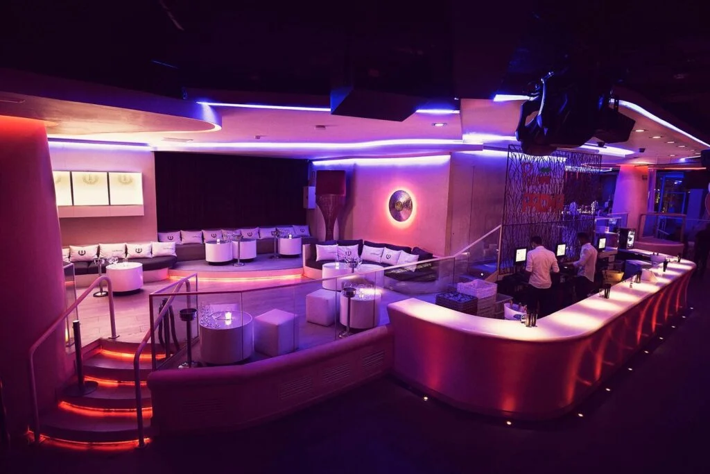 Interior de un club nocturno moderno en Barcelona con sofás blancos, barra iluminada y luces de neón en tonos rosa y azul