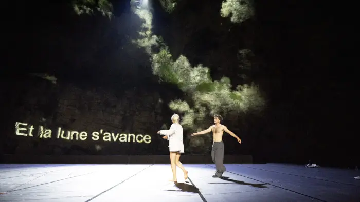Escena de una representación teatral contemporánea en un gran escenario oscuro, con dos intérpretes en movimiento, uno de ellos con el torso descubierto y el otro con una prenda clara, bajo una iluminación dramática. En el fondo se proyecta el texto “Et la lune s’avance” junto a una imagen atmosférica de nubes, creando un ambiente poético y experimental de danza y teatro físico..