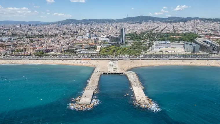 Vista aérea del Espigó del Gas en la playa de la Barceloneta, con el mar Mediterráneo en primer plano y el skyline de Barcelona, el Parc de la Ciutadella y la montaña de Collserola al fondo.