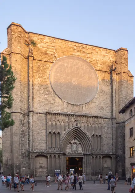 Facade of the Gothic Basilica of Santa Maria del Pi in Barcelona, featuring a large rose window and an ornate arched entrance, bathed in the warm glow of the setting sun. Visitors and tourists stroll through the historic square in front of the church.