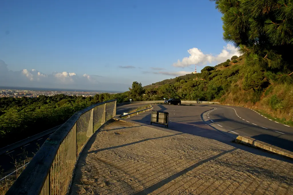 Mountain road leading to Sarrià Viewpoint in Barcelona, with a winding asphalt road, safety barrier and lush hillside vegetation, overlooking panoramic views of the city and the Mediterranean Sea beneath a clear blue sky.