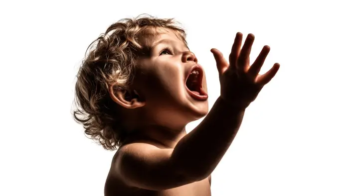 Retrato de un niño en estudio sobre fondo blanco, con expresión intensa y la boca abierta mientras levanta la mano hacia arriba, capturando un momento de emoción y energía..