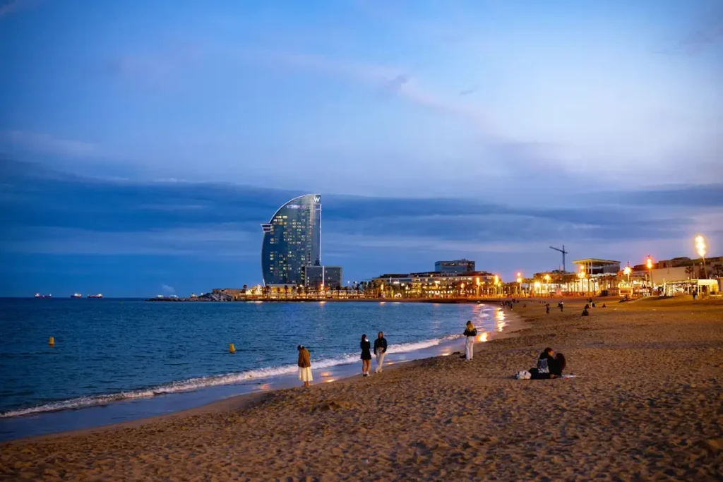 Vista de la playa de la Barceloneta al atardecer en Barcelona, con personas paseando junto al mar Mediterráneo y el emblemático Hotel W iluminado en el horizonte.