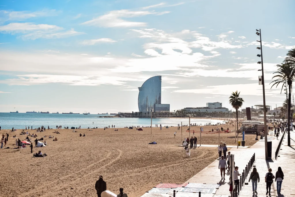 Vista panorámica de la playa de la Barceloneta en Barcelona con el Hotel W al fondo, paseo marítimo, palmeras y personas disfrutando de la costa mediterránea.