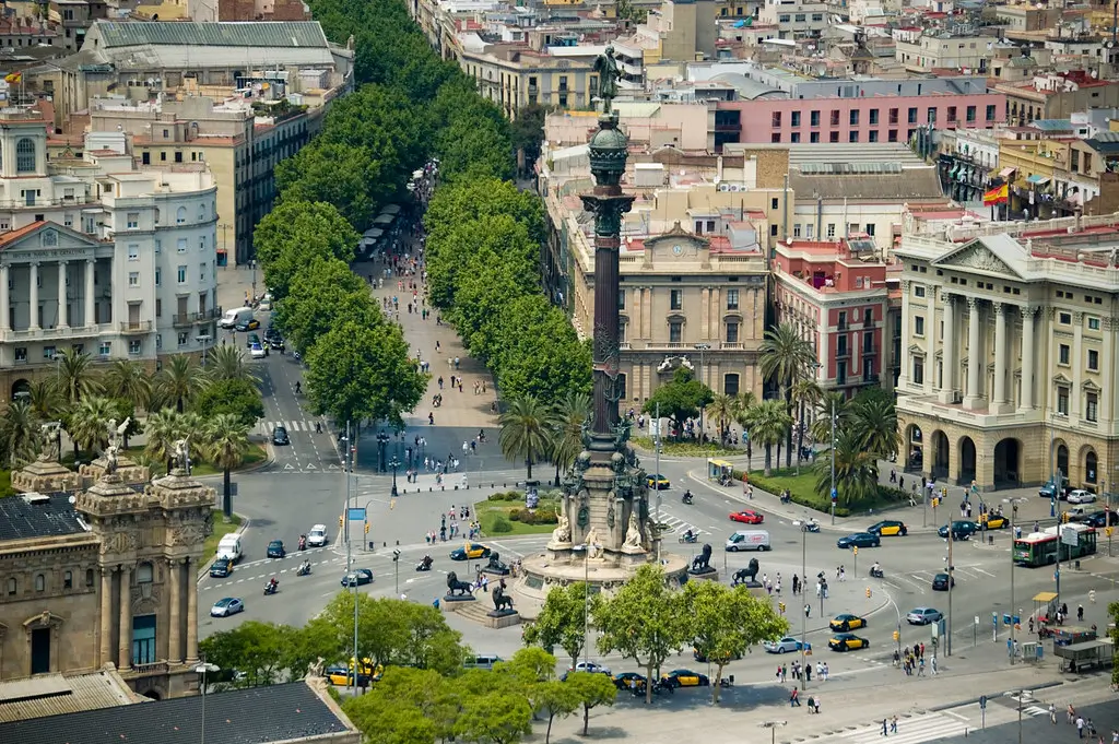 Aerial view of Plaça de Colom in Barcelona, featuring the Columbus Monument at the centre, surrounded by traffic roundabouts, palm trees and historic buildings, with the tree-lined La Rambla stretching into the heart of the city.