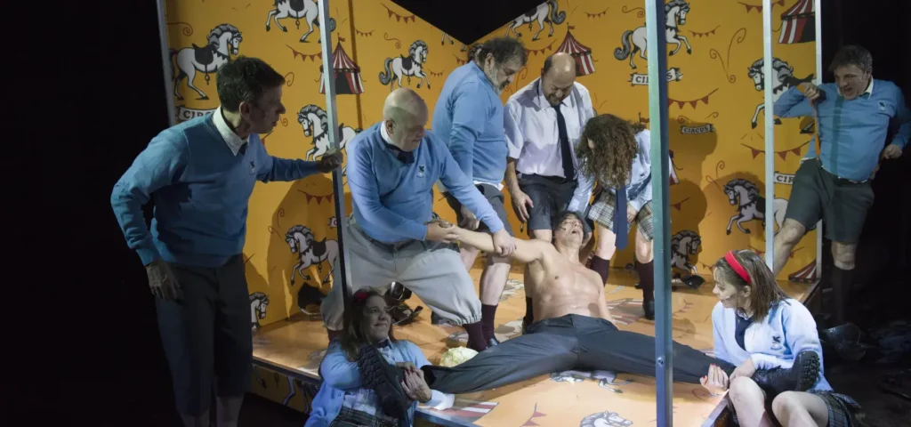 Escena de una representación teatral en la que varios personajes ayudan a un hombre semidesnudo en el centro del escenario, rodeado de otros actores vestidos con ropa escolar y deportiva, con un decorado de estilo circo amarillo de fondo con motivos de caballos y banderines, en una puesta en escena dramática inspirada en Ricardo III..