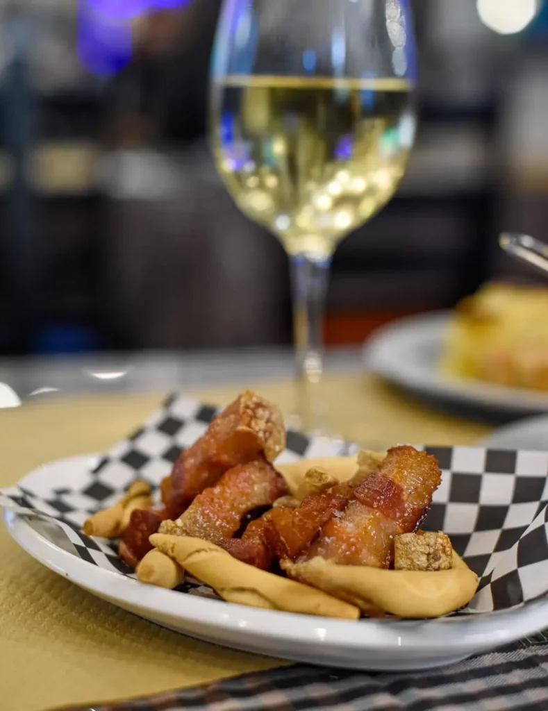 Plato de tapas tradicionales con panceta crujiente y aperitivos acompañado de una copa de vino blanco en un restaurante de La Barceloneta, reflejando la gastronomía local junto al mar en Barcelona.