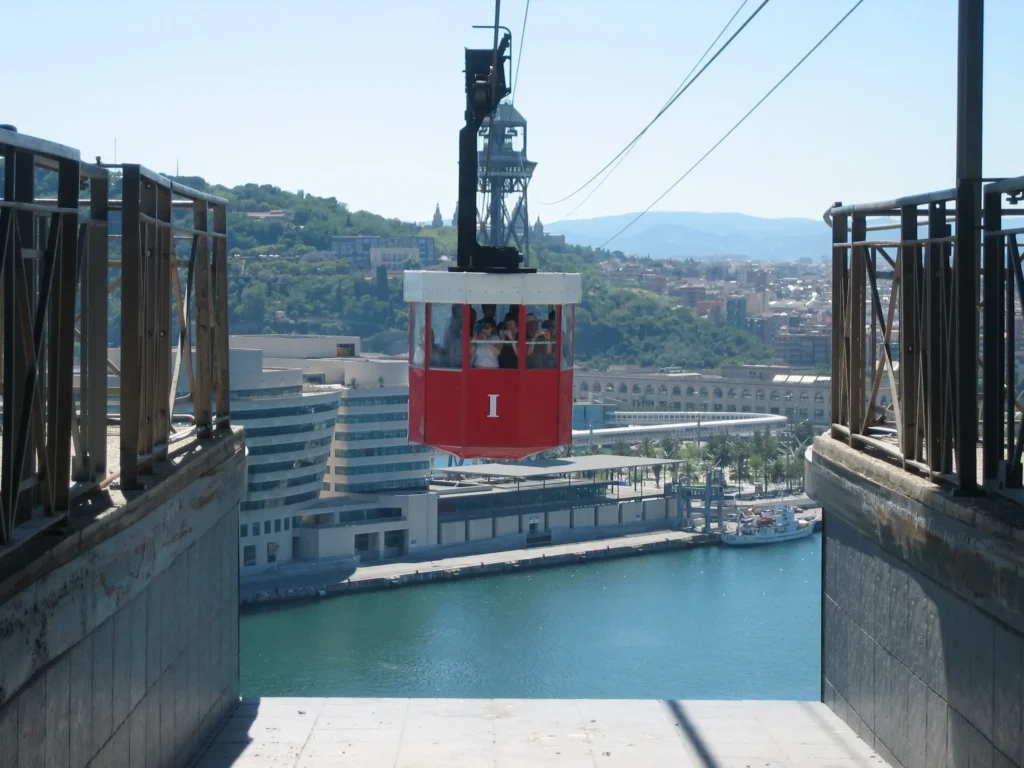 Red cabin of the Barcelona Port Cable Car crossing over Port Vell with passengers on board, suspended high above the water, with the city skyline and Montjuïc hill in the background on a clear sunny day.