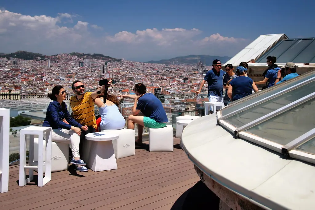 Group of people enjoying the rooftop terrace at MNAC in Barcelona, with panoramic views over the city, featuring high tables, a relaxing outdoor seating area, and a clear sunny sky.