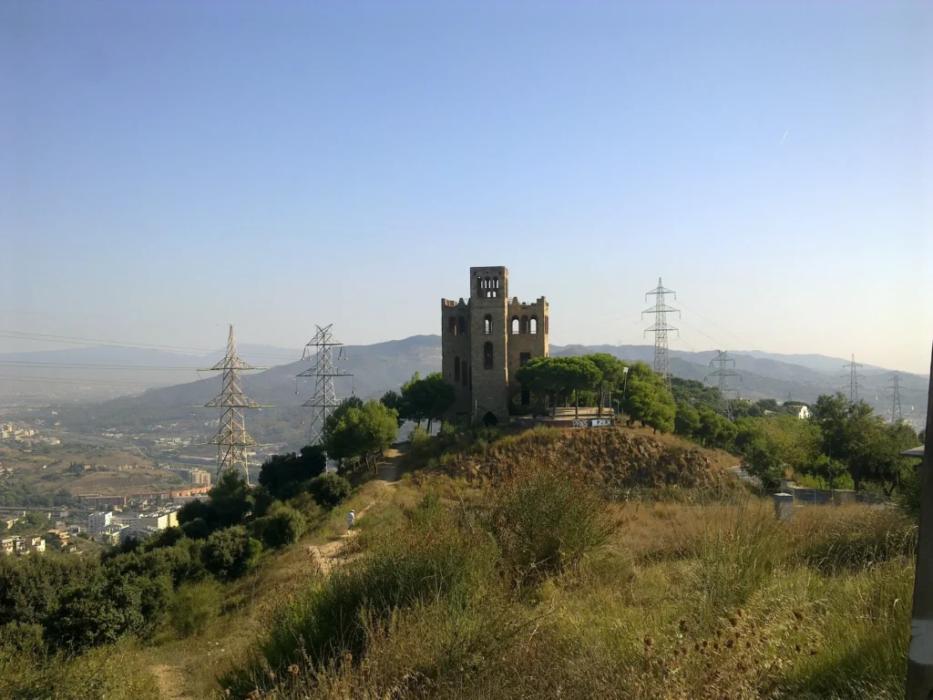 View of Torre Baró Castle in Barcelona, perched on a hill surrounded by lush greenery, walking trails and electricity pylons, with panoramic views of the city skyline and the surrounding mountains under a clear blue sky.