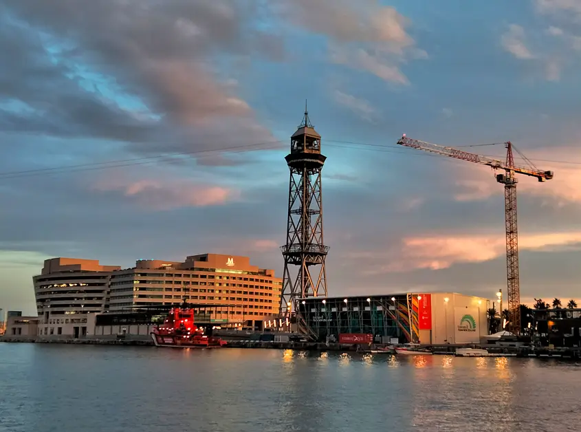 Vista de la Torre Jaume I junto al Port Vell de Barcelona al atardecer, con el World Trade Center, embarcaciones y reflejos sobre el agua bajo un cielo de tonos azules y anaranjados.