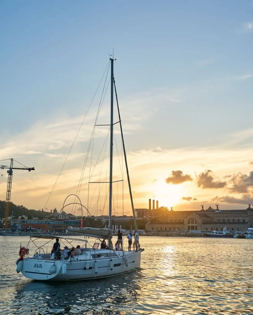 Velero navegando por las aguas del Port Vell de Barcelona durante el atardecer, con el sol reflejándose sobre el mar y edificios históricos del frente marítimo al fondo.