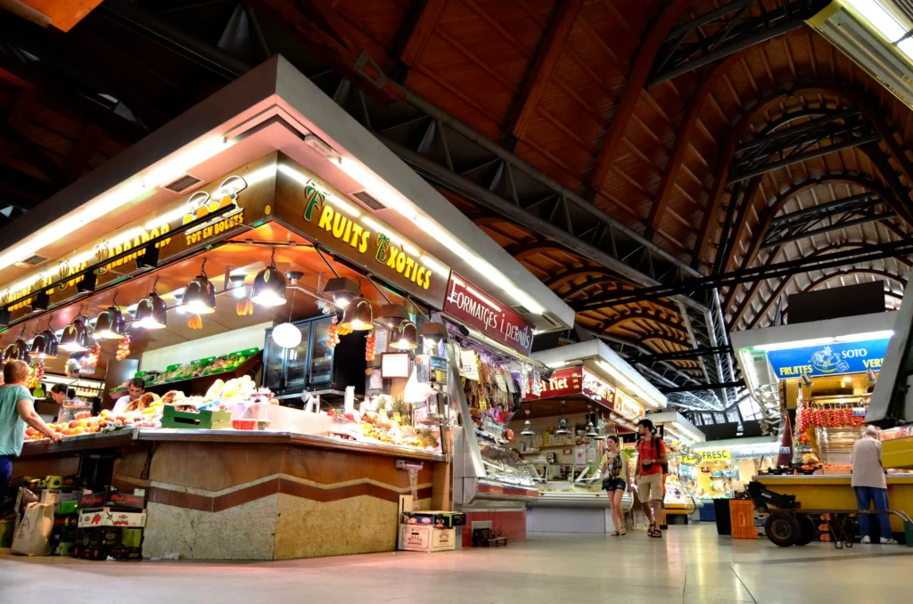 Interior of a traditional Barcelona market with fresh fruit, food stalls and shoppers beneath its distinctive wooden roof.