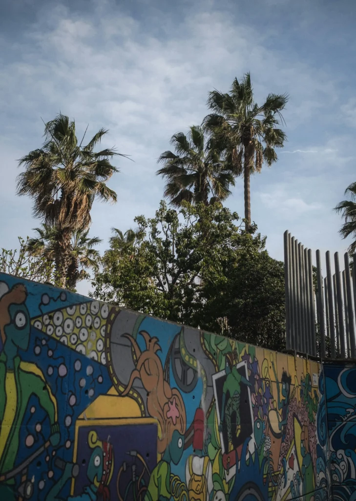 Colourful street art mural in Diagonal Mar, Barcelona, surrounded by palm trees and urban green spaces near the Parc del Fòrum waterfront.
