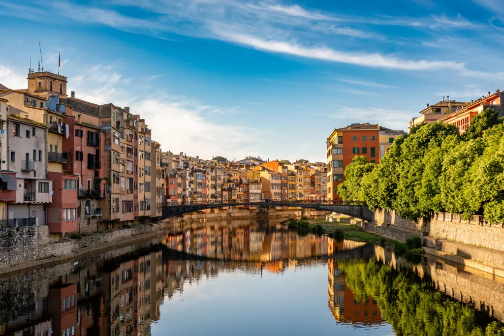 Farbenfrohe Häuser am Fluss Onyar in Girona mit ihren Spiegelungen im Wasser und einer Brücke im Hintergrund.