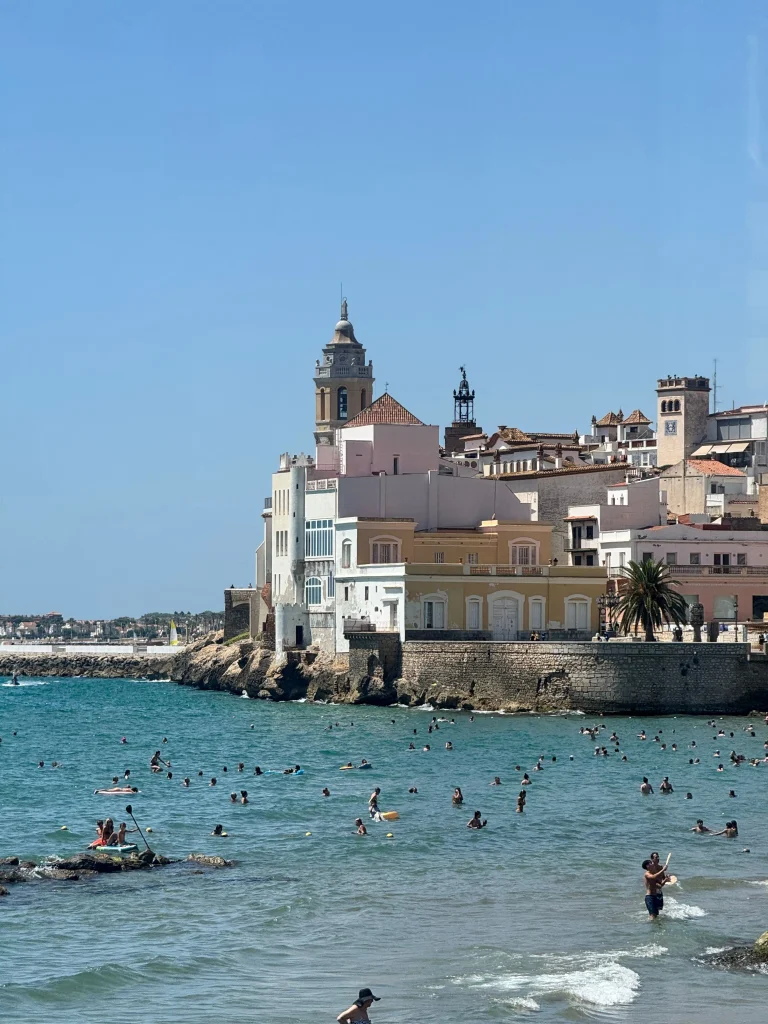 Blick auf den Strand von Sitges und die historische Strandpromenade mit Badegästen im Mittelmeer.