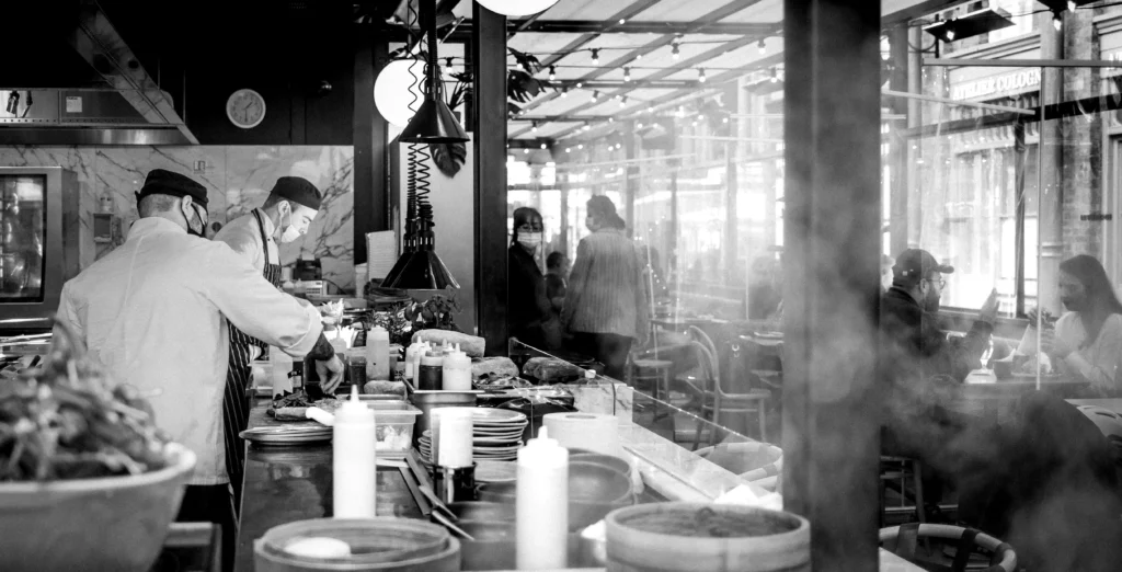 Dos cocineros preparan platos en una cocina abierta mientras los clientes disfrutan de su comida en el interior del restaurante.