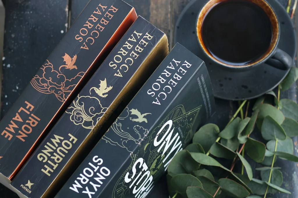 Tres libros de la autora Rebecca Yarros, titulados Iron Flame, Fourth Wing y Onyx Storm, colocados junto a una taza de café negro y hojas verdes sobre una mesa de madera.